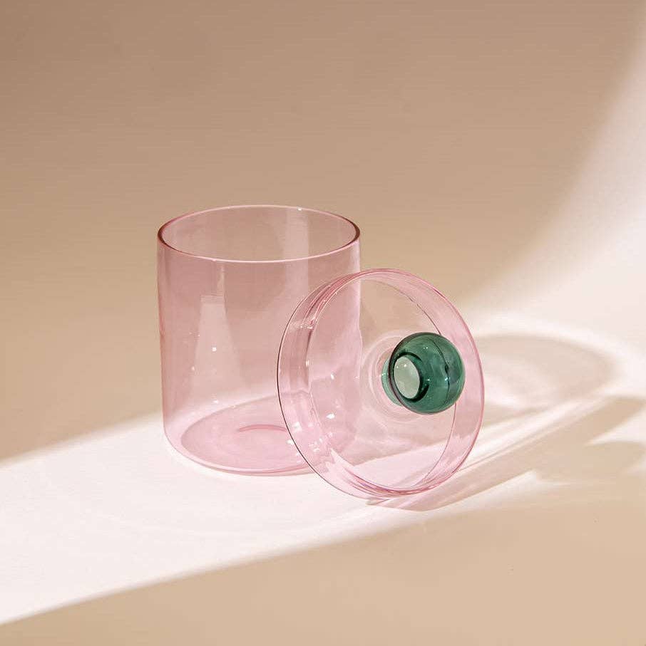 A translucent pink tumbler glass with its matching lid leaning against it on a warm beige surface, featuring a small green accent ring on the lid.