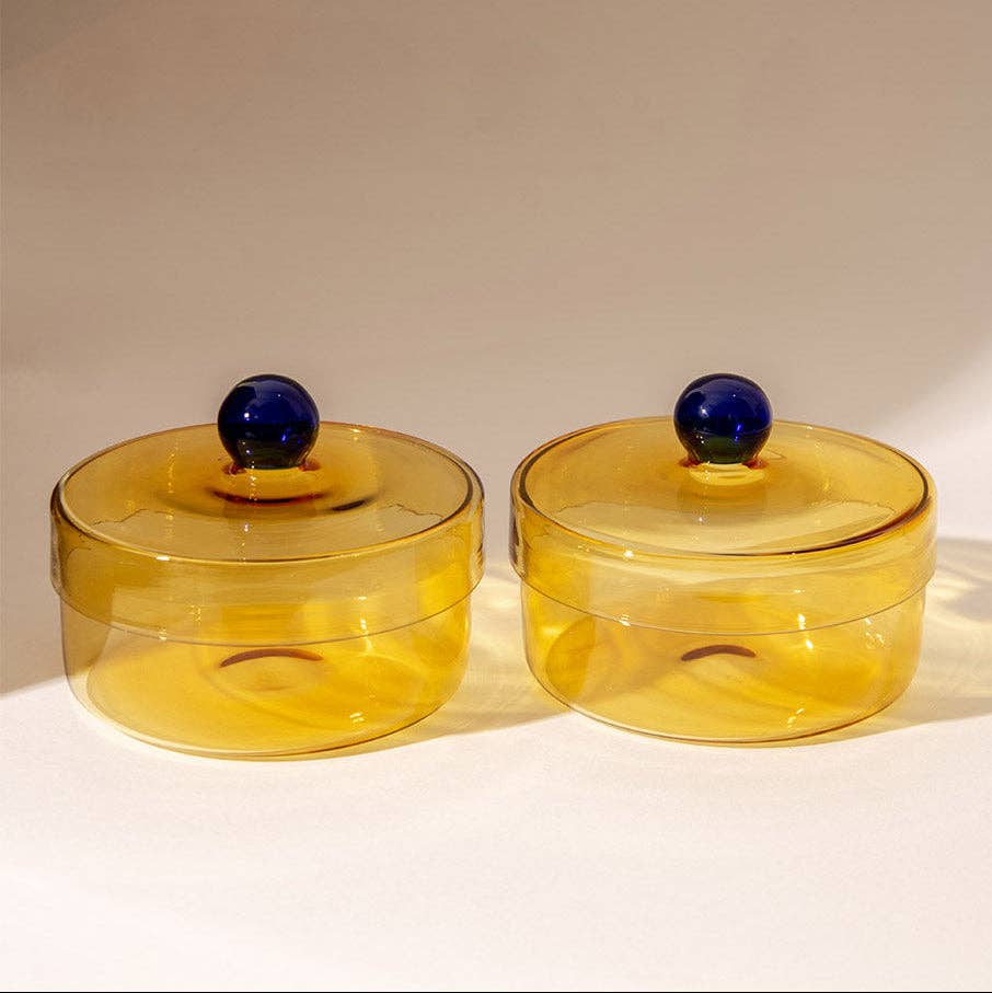 Two amber yellow translucent glass lidded boxes with dark blue spherical knobs, set against a plain light beige background.