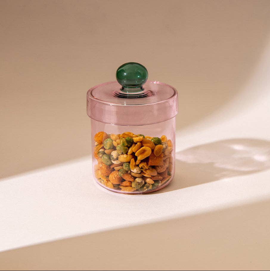 A translucent pink glass jar with a matching pink lid and a round green knob, filled with mixed nuts and dried fruit, set on a beige surface with soft shadows.