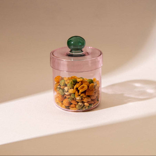 A translucent pink glass jar with a matching pink lid and a round green knob, filled with mixed nuts and dried fruit, set on a beige surface with soft shadows.