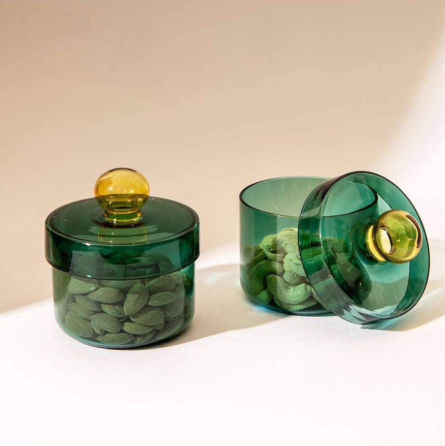 Two translucent dark green glass jars with round lids and gold-toned knobs, one closed and one open, both filled with green almonds on a warm beige background.