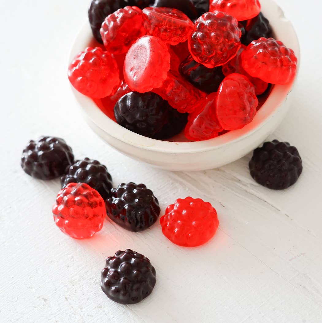 Gummy candies resembling red and black berries in a white bowl on a light surface.