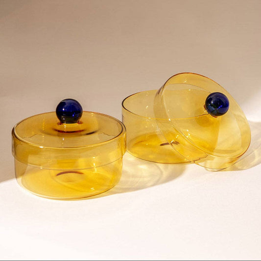 Two amber-yellow translucent glass lidded boxes with dark blue spherical knobs, displayed on a warm beige background.
