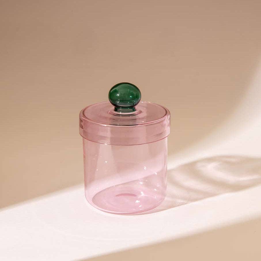 A translucent pink cylindrical container with a matching pink lid and a dark green spherical knob, set on a warm beige surface with soft shadows.