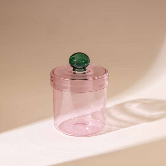 A translucent pink cylindrical container with a matching pink lid and a dark green spherical knob, set on a warm beige surface with soft shadows.