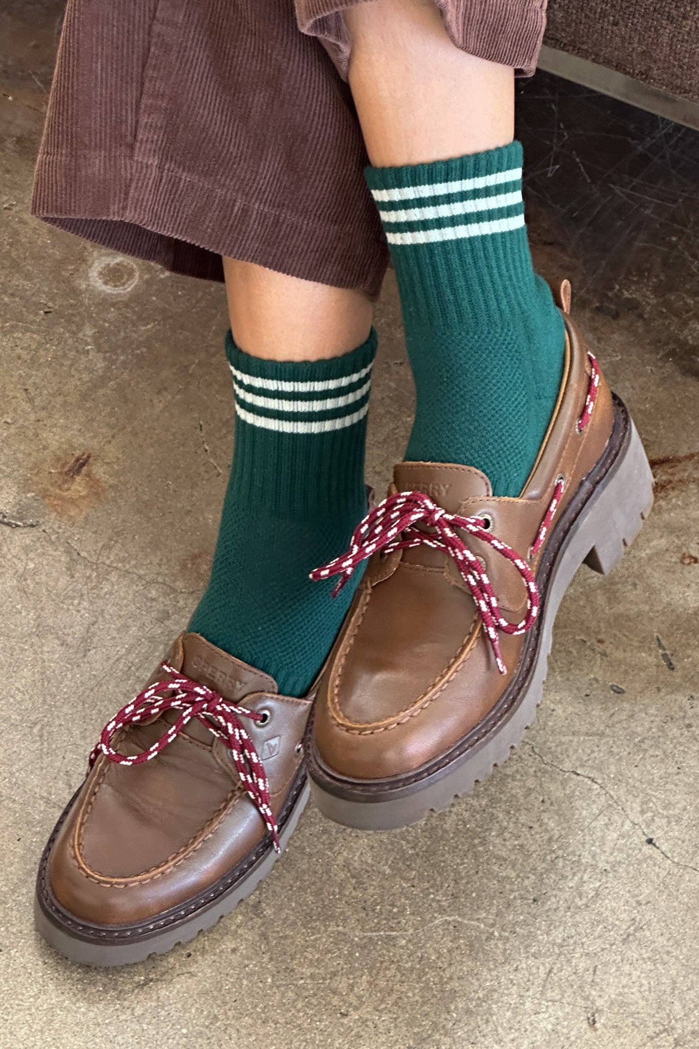 Brown loafers with red laces worn with green socks on a concrete floor.