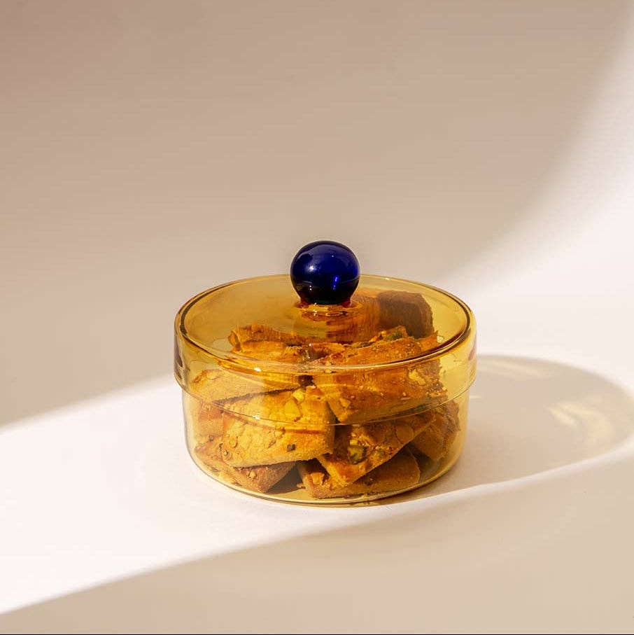 A round amber glass container with a clear lid and a dark blue spherical knob, filled with golden-brown chips, set on a white surface with warm sunlight and shadows.
