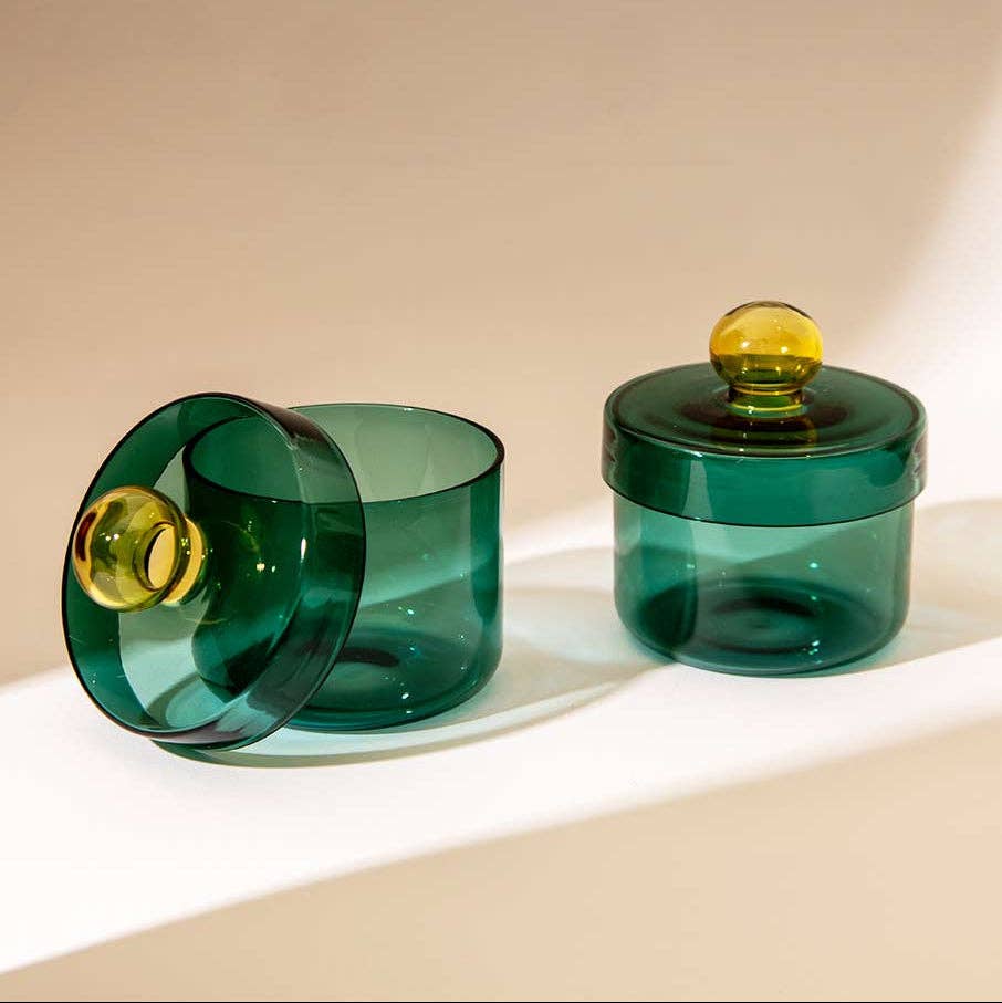 A pair of translucent dark green glass containers with matching lids, one standing upright and the other tipped on its side, both featuring round amber-yellow glass knobs, set against a warm beige background.