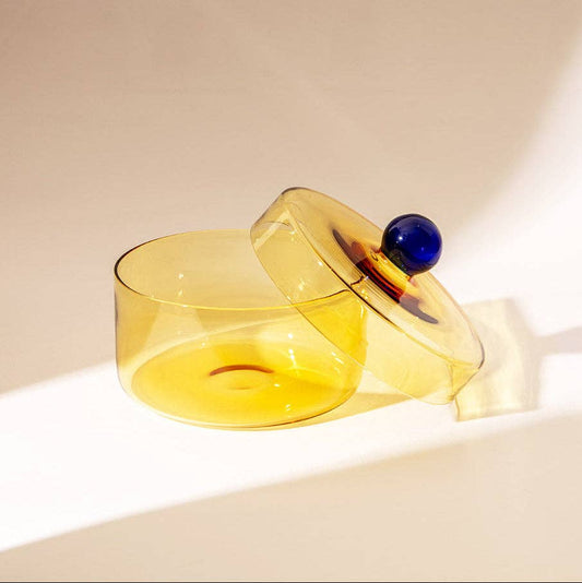 A translucent yellow glass container with a matching lid and a dark blue spherical knob, photographed on a warm off-white surface with strong sunlight and shadows.