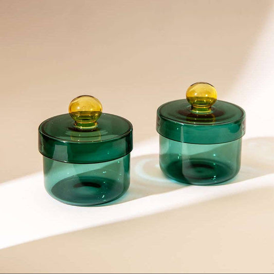 Two small translucent dark green glass jars with matching lids and round amber-yellow knobs, photographed on a warm beige surface with soft shadows.