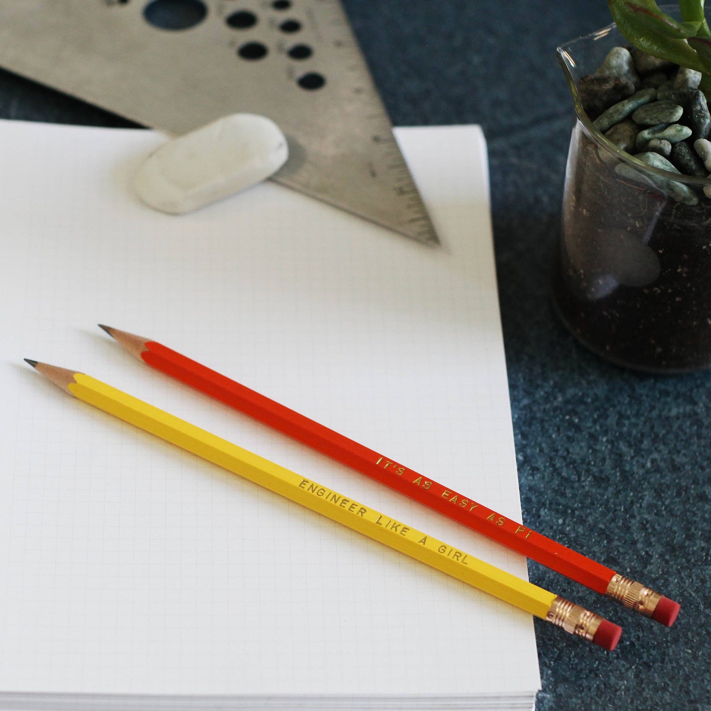 Two pencils, one yellow and one red, printed with the phrase 'ENGINEER LIKE A GIRL', resting on an open notebook alongside a metal ruler and eraser.