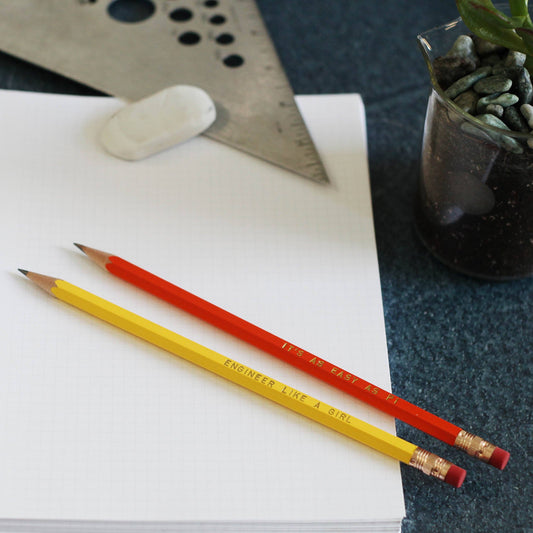 Two pencils, one yellow and one red, printed with the phrase 'ENGINEER LIKE A GIRL', resting on an open notebook alongside a metal ruler and eraser.