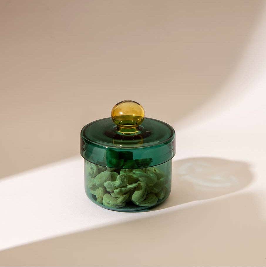 A small translucent dark green glass jar with a matching lid and a round amber-yellow knob, shown on a clean white surface with soft shadows.