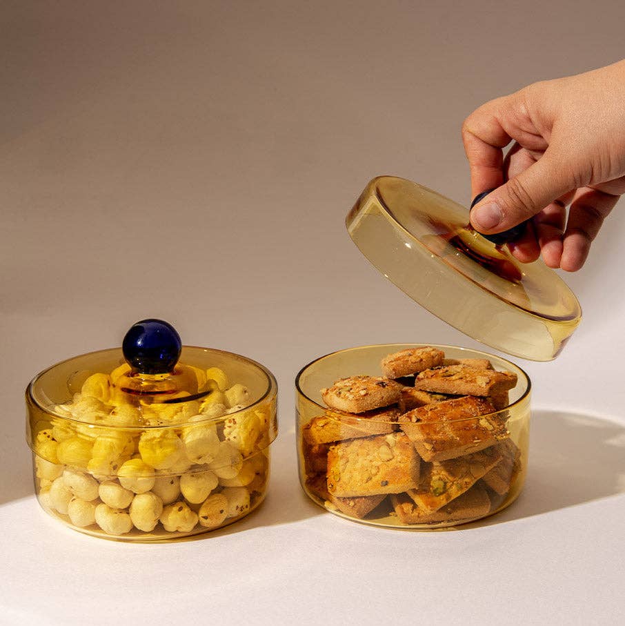 Two round translucent amber glass jars with lids, one filled with yellow marzipan and the other with cookies, on a beige background. A hand is lifting the lid of the right jar.