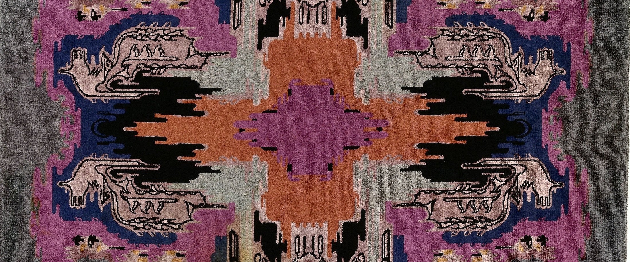 A square area rug with a symmetrical, geometric southwestern-inspired pattern in purple, orange, black, gray, and beige.