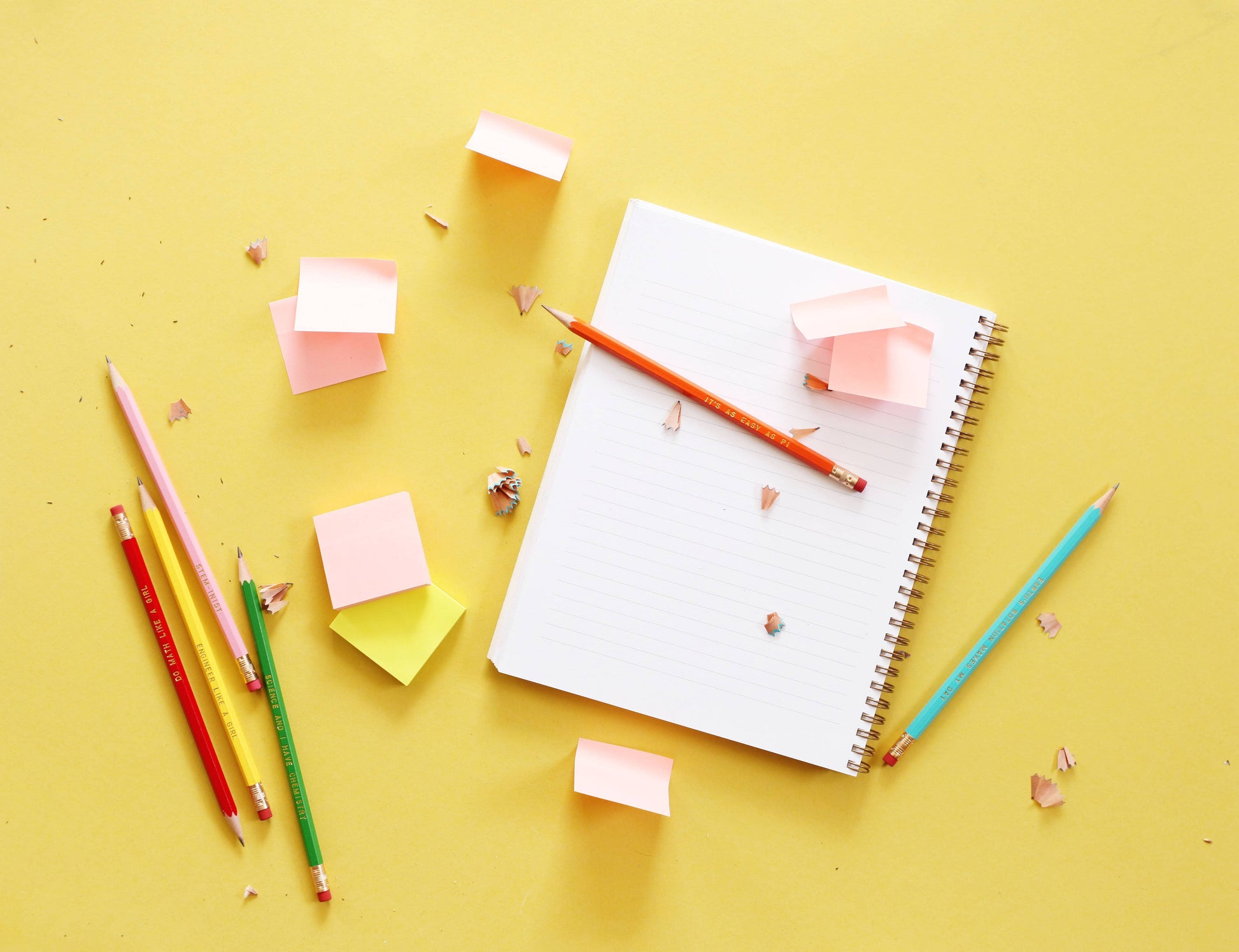 A spiral-bound lined notebook lies open on a bright yellow background, surrounded by colorful pencils, sticky notes, and small paper shavings. The notebook has an orange pencil resting across it, and additional colored pencils are placed to the left and right.